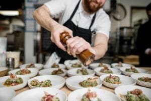 Transforma tu Cocina en una Máquina de Eficiencia - Bearded chef seasoning gourmet dishes with pepper in a busy restaurant kitchen.